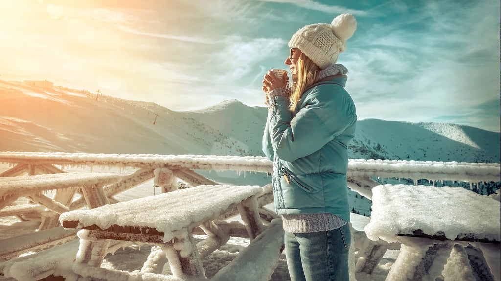 white woman sipping coffee wearing a snow hat and fleece, surrounded by snow covered tables overlooking the sun rise over a snowy mountain