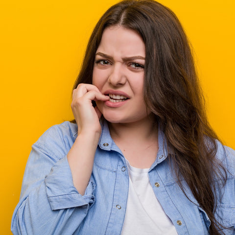 white woman with long brown hair in denim shirt with yellow background chewing her nail nervously