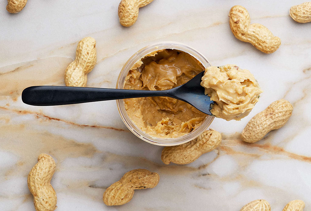 A black spoon, filled with peanut butter, lays on top of an open jar filled with peanut butter. Peanuts are scattered around the jar.