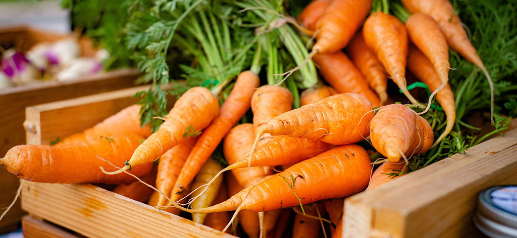 Freshly picked carrots lay in a wooden crate, with large leafy green roots.