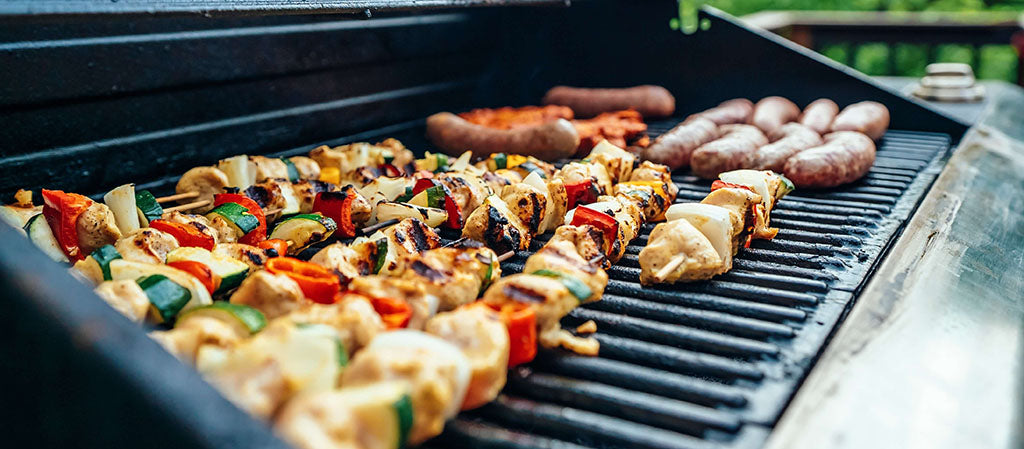 A close-up shot of a long rectangular BBQ which is ladened with chicken and vegetable skewers and sausages, which are charring well