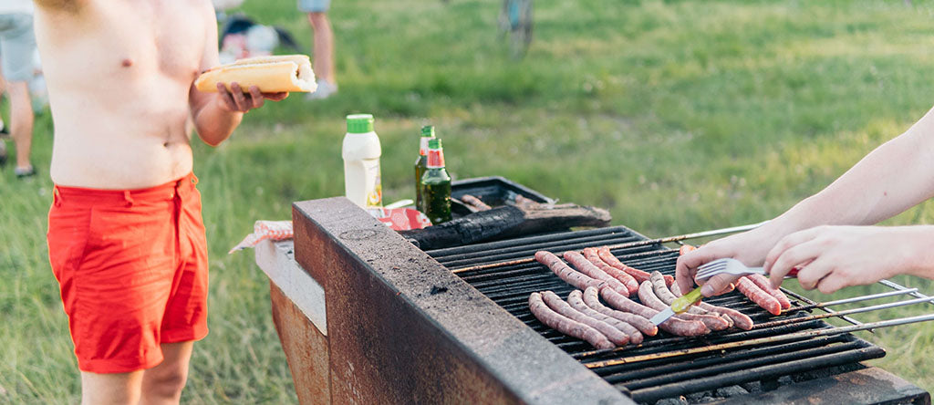 A cook-out is taking place (you can see a blurred collection of people stood on green grass in the background), and a pair of white male hands are flipping long hot dogs with a knife and fork. Another white male wearing glasses stands at the end of the BBQ in red shorts squeezing red sauce onto a hot dog. A pot of mayo and two beer bottles sit on the edge of the BBQ too