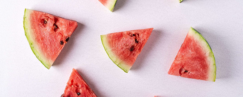 3 triangular slices of watermelon, with green rind and large, mature seeds lie spread out on light pink background.