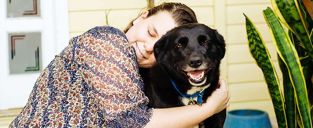 a white woman in a red and blue patterned top affectionately hugs her black dog that has their mouth open