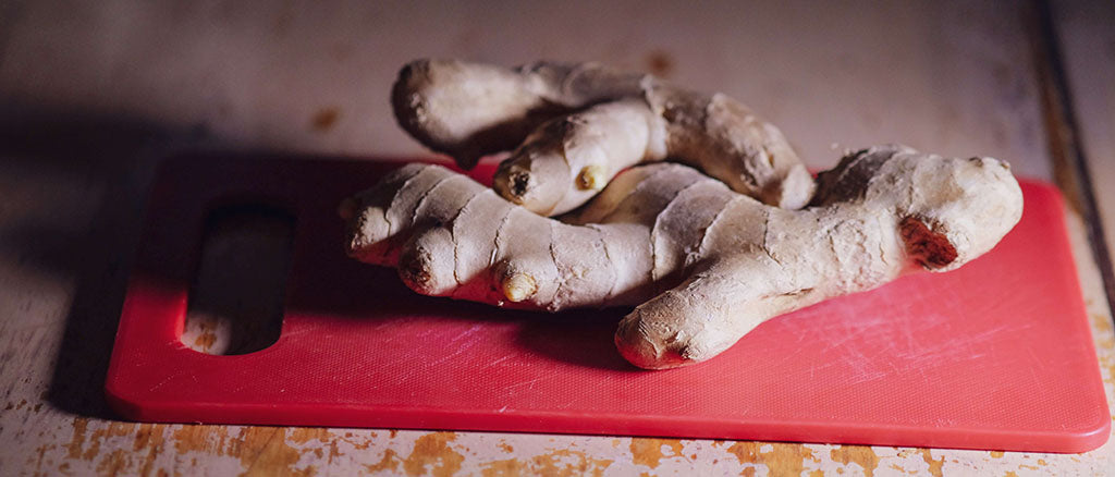 A hand of ginger root, with lots of off-shoot fingers, sits on top of a small, red, plastic chopping board