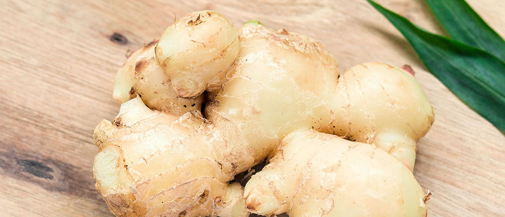 A hand of ginger root, sitting on a light wood chopping board with vibrant, dark green leaves across the top right-hand corner 