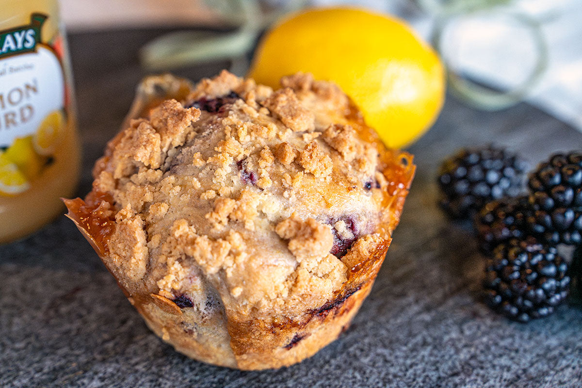A Blackberry Lemon Curd Muffin sits in front of blackberries, a lemon, and a jar of lemon curd