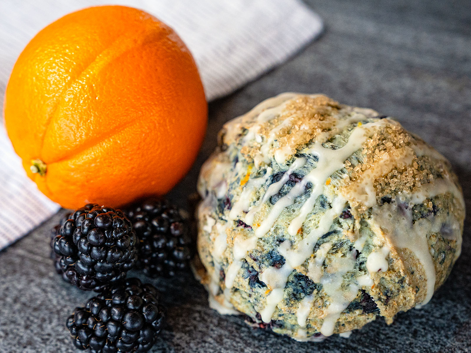 An orange and a pile of blackberries nestle beside an Orange Blackberry Scone