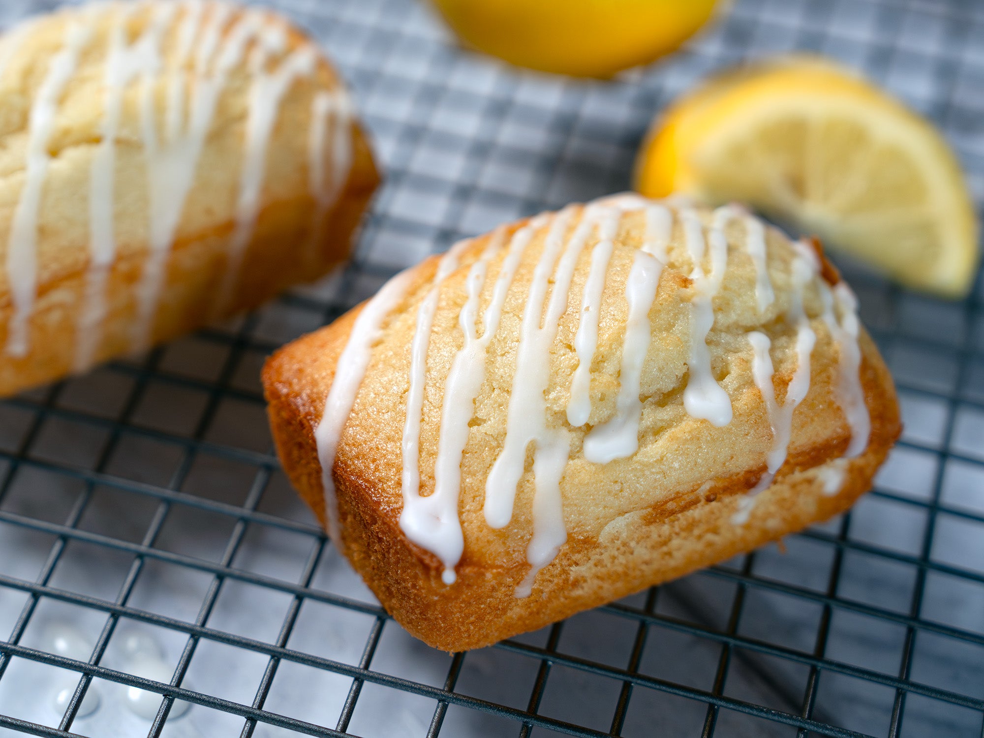 Two lemon mini loaves cool on a backing rack as a lemon rests behind them, out of focus.