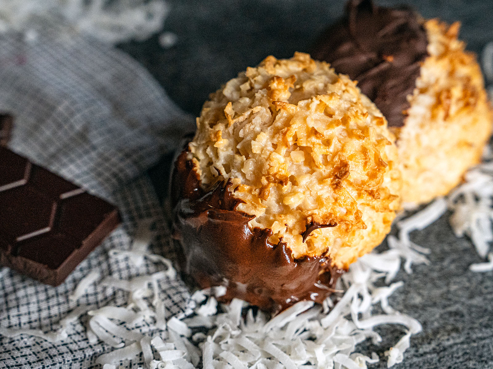 A freshly dipped coconut macaroon sits beside a chocolate bar, surrounded by coconut flakes