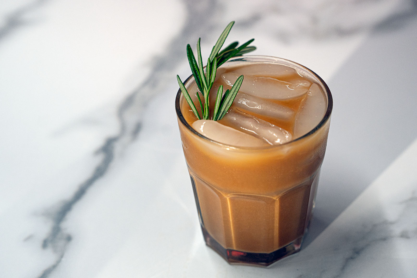 An iced rosemary brown sugar latte with a sprig of rosemary sits on marble