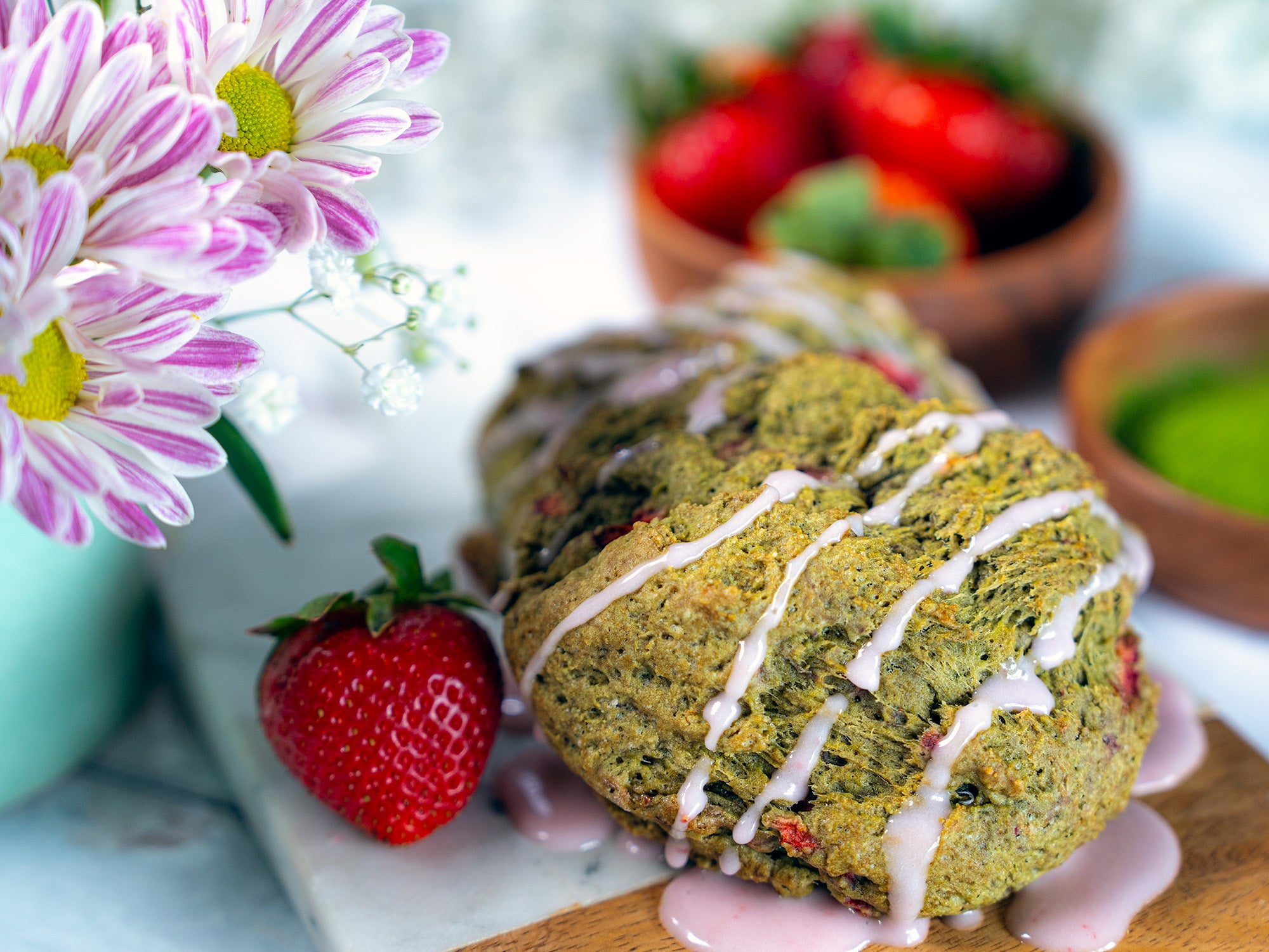 A freshly glazed scone drips with icing, beside a vase of purple flowers and a bowl of bright red strawberries.