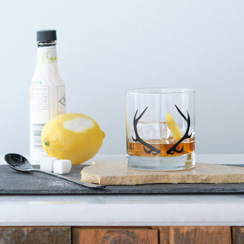 antler printed rocks glass with brown liquid and a lemon peel sitting next to spoon, lemon, and bitters bottle on stone table