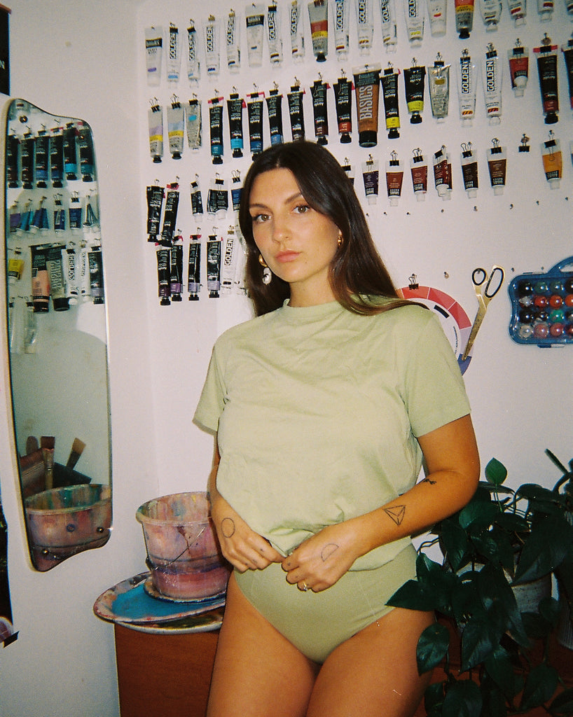 NICO at Home with Jess Cochrane. Jess stands in front of her wall of paints, wearing NICO Organic Bra  and Briefs in the colour Sage.