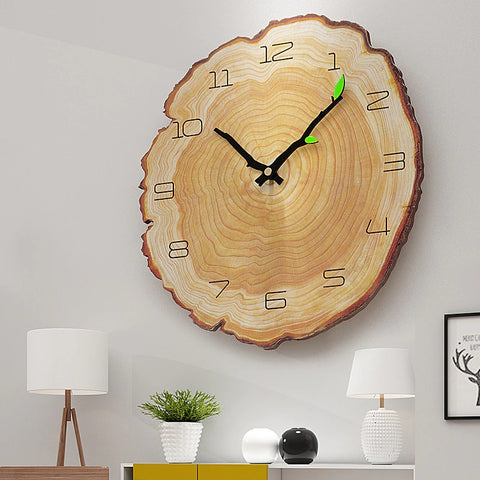A clock that is made out of a slice of wood complete with age rings within the wood slice. There are 2 black hands on the clock and each hand has a tiny green leaf on it. The clock is hanging on a wall with a table underneath with ornaments on it.