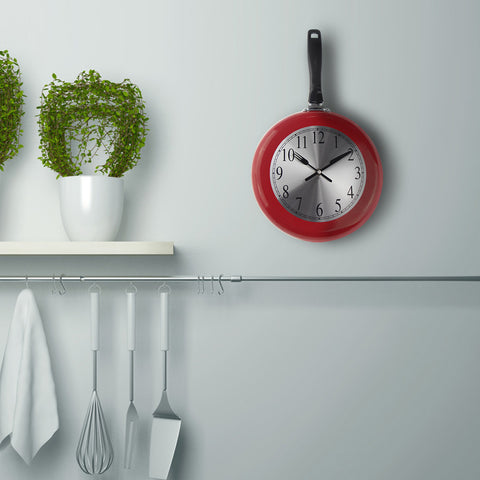A frying pan kitchen wall clock hanging on a kitchen wall. The clock is in the shape of a frying pan, the pan is red and it has a black handle. The hands are a fork and knife which reads 10:10am. There are some kitchen utensils hanging underneath as well as some small fake plants in pots