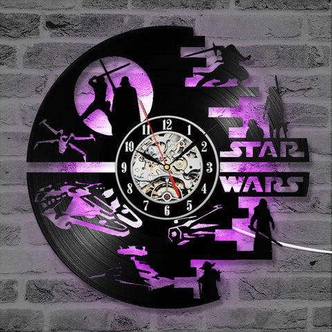 A Star Wars wall clock hanging on a greyish white brick wall.. The clock itself is a black vinyl record, and carved out of one side are silhouettes from the Star Wars world. It also has a purple backlight which lights up the entire clock.