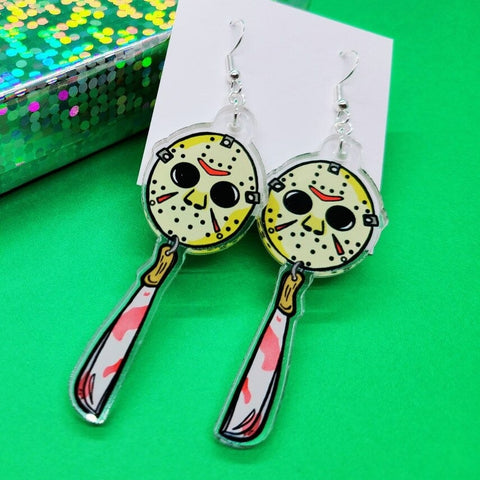 A pair of earrings inspired by the movie Friday the 13th. Each earring features Jason Voorhees hockey mask and hanging under the mask is a blood stained machete. The earrings are resting on a green background.
