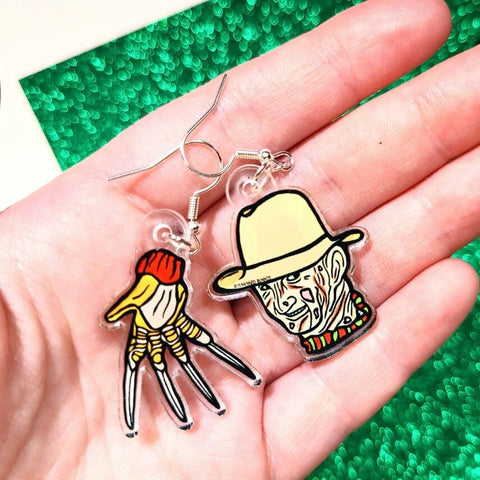 A pair of earrings inspired by the movie A Nightmare on Elm Street. One earring features Freddy Krueger's face and the other is of his glove made from knives. The earrings are in the palm of a persons hand.