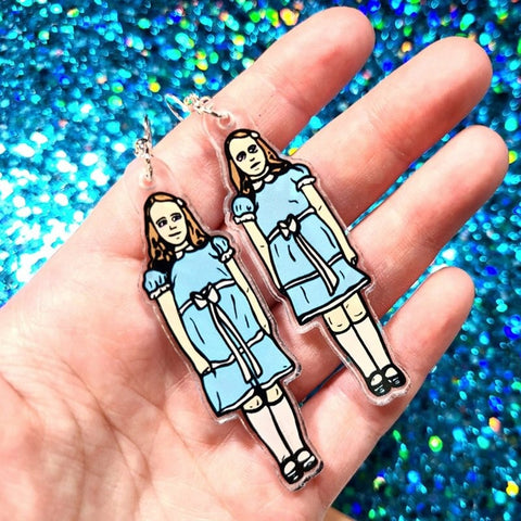 A pair of earrings inspired the creepy twin girls in the movie The Shining. One twin is on each of the earrings and they are wearing the same blue dress and white sash that they wore in the movie. The earrings are in the palm of a hand showing them close-up.