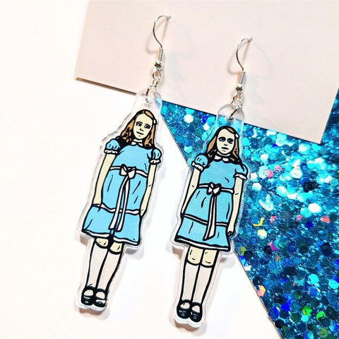 A pair of earrings inspired the creepy twin girls in the movie The Shining. One twin is on each of the earrings and they are wearing the same blue dress and white sash that they wore in the movie. The earrings are resting on a white and blue background.