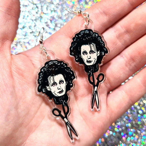 A pair of earrings inspired the movie Edward Scissorhands. Each earring features the face of Edward Scissorhands with a pair of scissors hanging underneath his head. The earrings are in the palm of a hand showing them close-up.