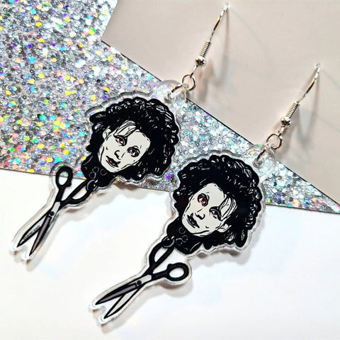 A pair of earrings inspired the movie Edward Scissorhands. Each earring features the face of Edward Scissorhands with a pair of scissors hanging underneath his head. The earrings are on a white and glittery background.