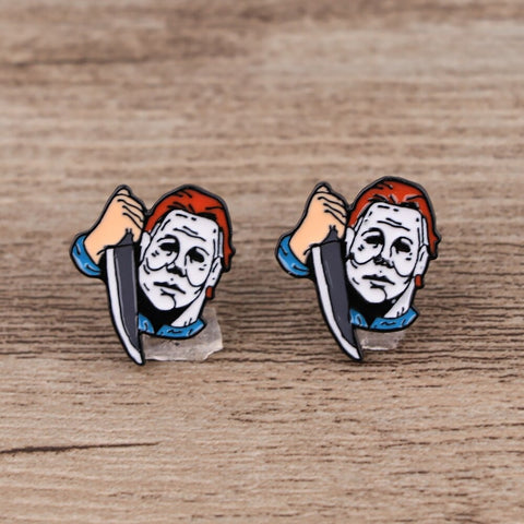 A pair of stud earrings inspired by the movie Halloween and the character Michael Myers. Each earring features the creepy white mask that Michael Myers wears and he is holding a knife. The earrings are on a wooden background.