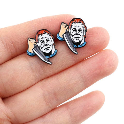 A pair of stud earrings inspired by the movie Halloween and the character Michael Myers. Each earring features the creepy white mask that Michael Myers wears and he is holding a knife. The earrings are in the palm of a hand showing them close-up.
