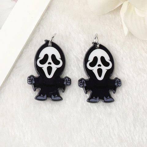 A pair of stud earrings inspired by the movie franchise called Scream. Each earring features the character called Ghostface who wears a black cloak and a terrifying white mask. The earrings are on a cream-coloured background.