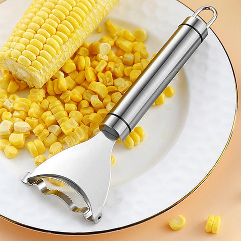 A silver metal kitchen corn stripper resting on a plate which has a cob of corn and loose corn kernels after being stripped off the cob.