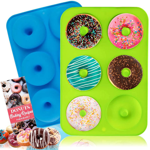 Two silicone donut molds, one blue and one green. There are 6 donuts in the green mold and the blue mold is reversed. There are cooked donuts and a recipe book nearby.