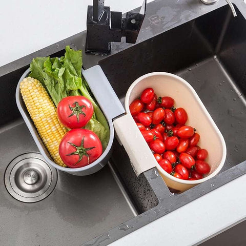 One grey and one white fruit and vegetable washer. Both washers are attached to a kitchen sink and have freshly washed vegetables in them.