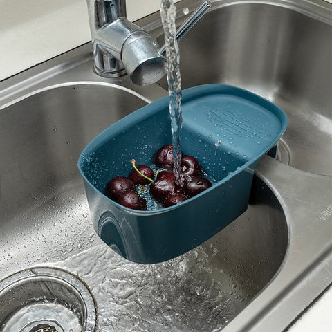 A dark blue fruit and vegetable washer attached to a kitchen sink. Fresh water is coming out of the facet and washing cherries which are inside the container.