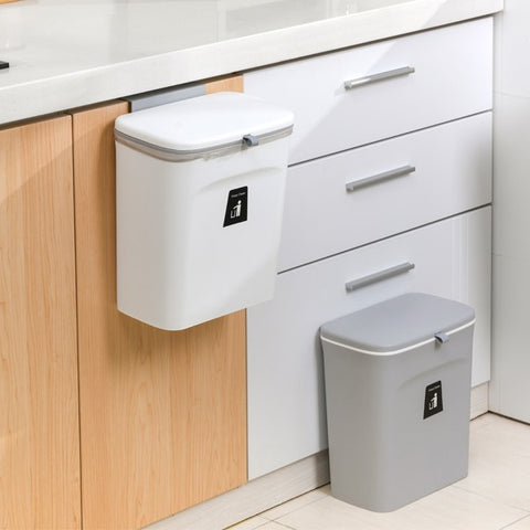 A small compact grey trash can on a kitchen floor along with a white compact trash can attached to a kitchen cupboard door.