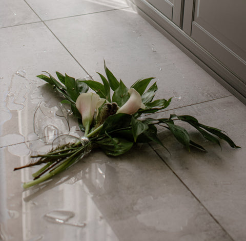 A broken glass vase on a white tiled floor with long stemmed scattered white flowers around it.