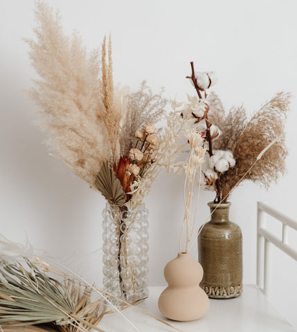 A glass vase alongside two ceramic vases. The 3 vases are filled with a variety of dried flowers.