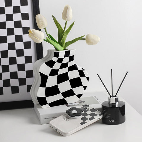 A vase with white tulips on a table. The vase has a black and white checkerboard design on it. Also on the table is a reed diffuser, cell phone, and a checked photo leaning against the back wall.