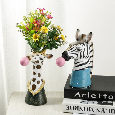 A giraffe shaped vase and a zebra shaped vase. The giraffe vase has a bouquet of colorful flowers in it and is white with black spots. It also has a pink chewing gum bubble coming from its mouth. The zebra vase is black and white striped and also has a pink chewing gum bubble coming from its mouth.