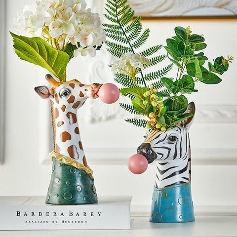 A giraffe shaped vase and a zebra shaped vase. The giraffe vase has white flowers and a large green leaf in it and is white with orange spots. It also has a pink chewing gum bubble coming from its mouth. The zebra vase has a bouquet of large leaves and is back and white striped and also has a pink chewing gum bubble coming from its mouth.