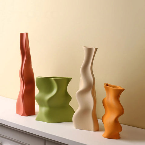 Four wavy shaped vase. 2 tall vases, one is burnt orange the other is cream. Plus 2 small vases, one is green the other is bright orange. All the vases are on a white shelf.