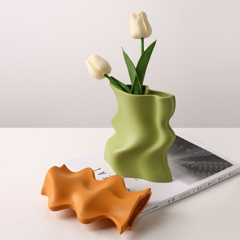 Two wavy shaped small sized vases. One is green and has 2 white tulips in it, the other is bright orange. The vases are on a white shelf with the green vase resting on a flat book.