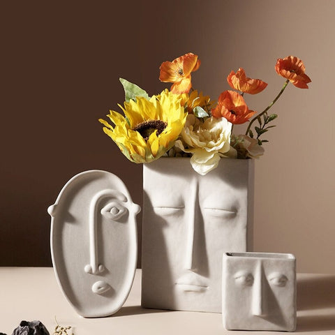 3 cream colored vases with abstract faces on them in 3 different sizes and shapes. The largest vase has a bouquet of yellow and orange faux flowers in it and the other 2 are empty.