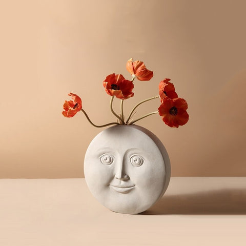 A cream colored circular vase which features an abstract shaped face on it. There are 5 burnt orange faux poppy flowers in the vase.