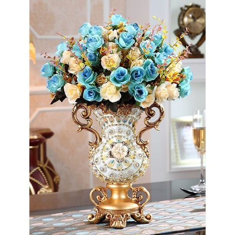 A lavish white and gold antique-looking vase with a floral design in the centre and gold twin handles on the side. The vase is filled with a giant bouquet of blue and cream colored roses.