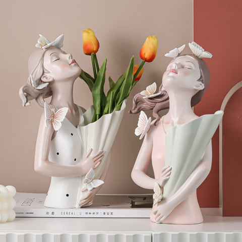 Two ceramic vases in the shape of girls from the waist up. The girls are surrounded with butterflies and their heads and noses are pointing upwards into the air and they are also holding vases which are actual real vases to hold flowers. One of the vases has 3 orange tulips in it.