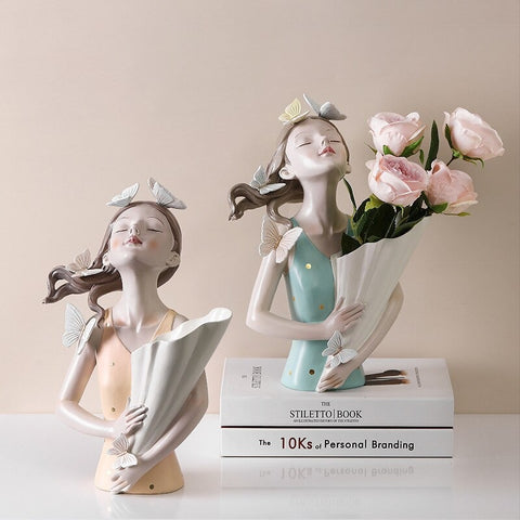 Two ceramic vases in the shape of girls from the waist up. The girls are surrounded with butterflies and their heads and noses are pointing upwards into the air. Both vase are on a table and one vase is resting on a book and has four pink roses in it.