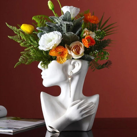 A white vase in the shape of a woman’s head and shoulders portrait. The head is cut off above the nose which is where flowers can be placed. There is a large bouquet of colorful flowers in the vase.