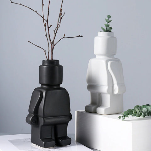 Two giant Lego man vases with holes in the head for flowers. The vases are shaped like Lego man and are black and white. One vase has a decorative branch in it and the other has a green leafy plant in it.