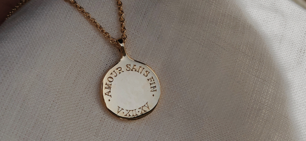 A gold pendant necklace with the words "Endless Love end" in French and a date styled in Roman numerals hand engraved.